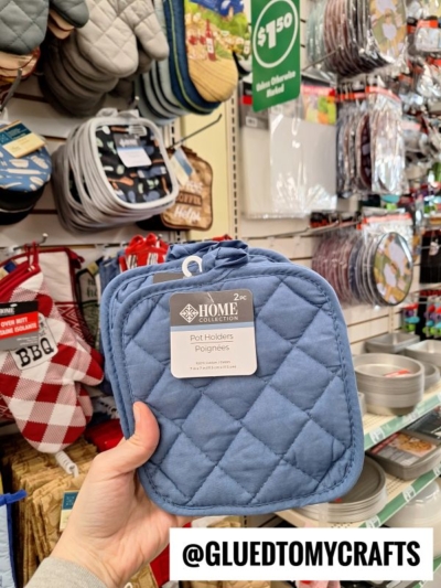 A hand holds up a pack of two blue quilted pot holders in a store aisle filled with various kitchen items and accessories. The image features the Instagram handle @GLUEDTOMYCRAFTS.