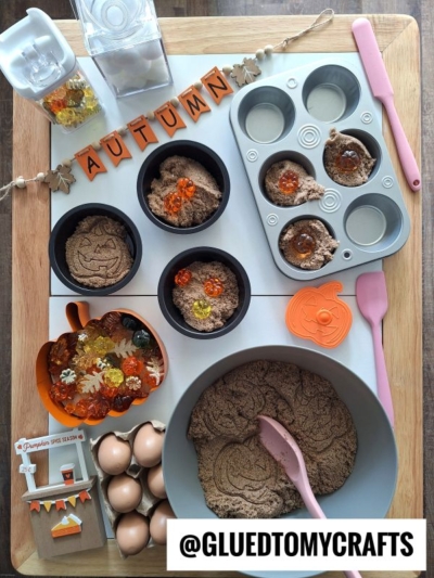 A flat lay of autumn-themed sensory play items: muffin tins with brown sensory sand, pumpkin and gem decorations, play eggs, a bowl with a pink spoon—perfect for kinetic sand play ideas—plus an autumn banner and a sign reading “@GLUEDTOMYCRAFTS.”.