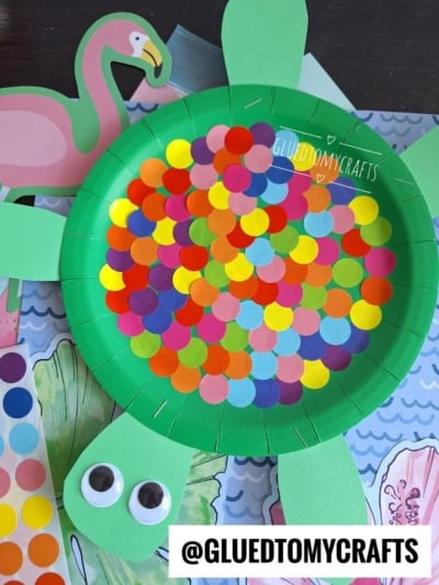 A green paper plate turtle craft with colorful paper dots on its shell, green paper legs and head, and two large googly eyes, displayed on a decorated background with a pink flamingo illustration.