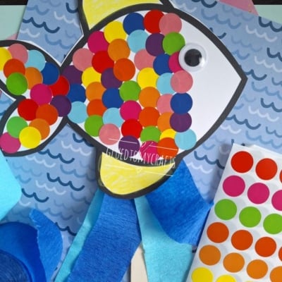 A paper fish craft decorated with colorful round stickers and a googly eye is placed on a blue wavy background. Craft materials, including blue crepe paper and a sticker sheet, are visible. @GLUEDTOMYCRAFTS text is at the bottom.
