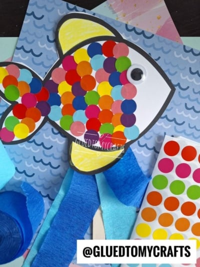 A paper fish craft decorated with colorful round stickers and a googly eye is placed on a blue wavy background. Craft materials, including blue crepe paper and a sticker sheet, are visible. @GLUEDTOMYCRAFTS text is at the bottom.