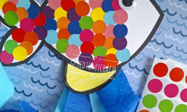A paper fish craft decorated with colorful round stickers and a googly eye is placed on a blue wavy background. Craft materials, including blue crepe paper and a sticker sheet, are visible. @GLUEDTOMYCRAFTS text is at the bottom.