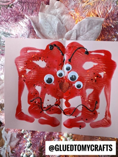 A red, symmetrical paint splat love monster butterfly with googly eyes, a heart-shaped nose, and black glitter sits in front of a pink Christmas tree. @GLUEDTOMYCRAFTS is written at the bottom.