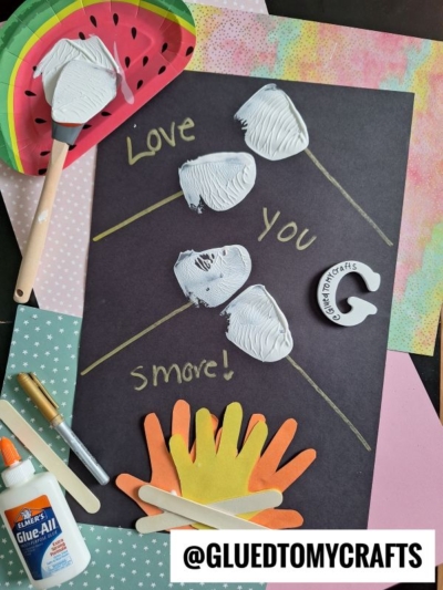 A spatula stamped smores craft on black paper features three white handprints forming hearts, gold lines connecting them, and the words Love You Smore! Orange and yellow paper hands, craft sticks, glue, and a painted watermelon are included.