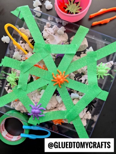 A sensory bin with play sand, plastic spiders, and small toys is covered with green tape arranged like a spiderweb. Perfect for kinetic sand play ideas! Tweezers, tape, and toy bats are nearby. The handle @GLUEDTOMYCRAFTS is at the bottom.