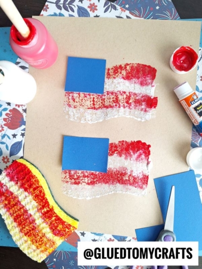 A craft setup showing two American flag art pieces made with blue paper squares and red and white sponge-painted stripes, surrounded by paint, a sponge, scissors, glue, and floral paper. @GluedToMyCrafts is written at the bottom.