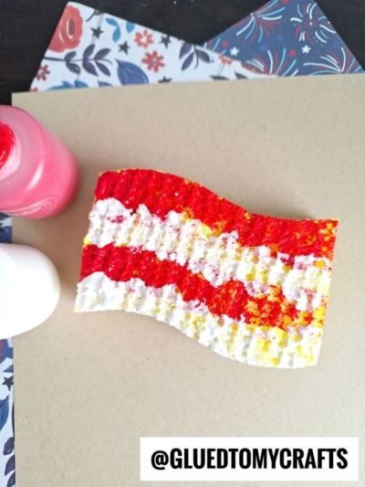 A wavy piece of cardboard painted with red, yellow, and white stripes lies on a tan surface next to bottles of pink and white paint, with a corner of floral-patterned paper visible.
