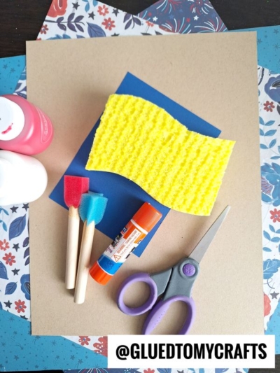 A yellow sponge, blue and red foam brushes, glue stick, scissors, and pink and white paint bottles on top of layered floral and plain colored paper. @GLUETOMYCRAFTS is written at the bottom.
