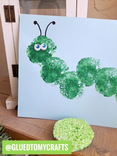 A HUNGRY CATERPILLAR CRAFT made from green sponge paint prints on blue paper, with googly eyes and drawn antennae. A round green sponge sits in front. @GLUETOMYCRAFTS is labeled at the bottom.