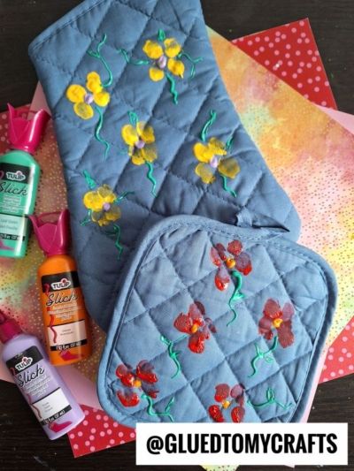 Two quilted blue pot holders with hand-painted yellow and red flowers are displayed on patterned paper backgrounds. Bottles of fabric paint are placed beside them. A text label reads @GLUEDTOMYCRAFTS.