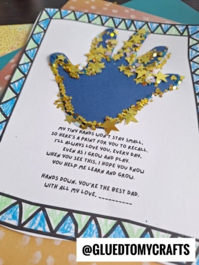 A handmade card features a blue handprint outlined with gold star-shaped glitter. Below the handprint is a heartfelt poem about a childs hands growing. The card is decorated with colorful patterns and @GLUEDTOMYCRAFTS is at the bottom.