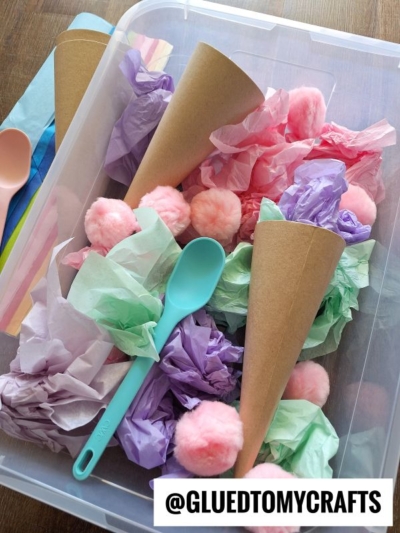 A plastic bin filled with brown paper cones, pastel tissue paper, pink pom-poms, and a blue plastic spoon creates a fun tissue paper ice cream sensory bin. The @GLUEDTOMYCRAFTS username appears on a label in the bottom right corner.