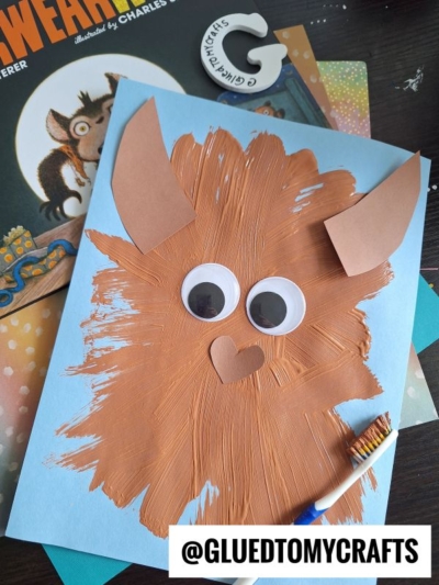 A blue sheet of paper painted brown with a toothbrush to resemble a monster, featuring large googly eyes, paper ears, and a heart-shaped nose, alongside a book and crafting supplies.