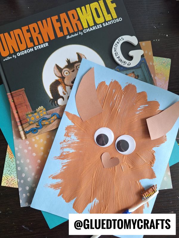 A children’s book titled UNDERWEARWOLF is shown with a handmade brown wolf craft featuring googly eyes on blue paper. Various colored papers are beneath the book and craft. Text reads @GLUEDTOMYCRAFTS.
