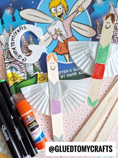 Craft supplies and materials for making fairy puppets, including markers, a glue stick, wooden sticks with drawn faces and colored outfits, paper cupcake liners for wings, and a craft blog handle @gluedtomycrafts.