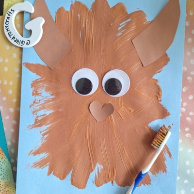 A brown, furry monster made with paint and paper pieces is glued onto blue paper. The monster has big googly eyes, pointy ears, and a heart-shaped nose. A toothbrush with brown paint lies next to the artwork.