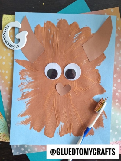 A brown, furry monster made with paint and paper pieces is glued onto blue paper. The monster has big googly eyes, pointy ears, and a heart-shaped nose. A toothbrush with brown paint lies next to the artwork.