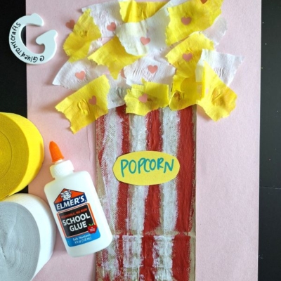 A crepe paper popcorn craft for kids featuring a popcorn tub made with red and white paint, tissue and crepe paper popcorn, a glue bottle, and craft supplies on pink paper. The word popcorn is written on a yellow oval.