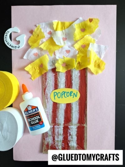 A crepe paper popcorn craft for kids featuring a popcorn tub made with red and white paint, tissue and crepe paper popcorn, a glue bottle, and craft supplies on pink paper. The word popcorn is written on a yellow oval.