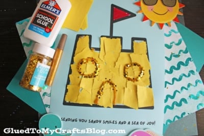 A handmade sandcastle art project made with torn yellow paper and gold glitter on blue paper, with a sun wearing sunglasses in the corner. Elmers glue, glitter, and a craft pen are nearby. Text reads, Sending you sandy smiles and a sea of joy!.