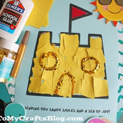 A handmade sandcastle art project made with torn yellow paper and gold glitter on blue paper, with a sun wearing sunglasses in the corner. Elmers glue, glitter, and a craft pen are nearby. Text reads, Sending you sandy smiles and a sea of joy!.