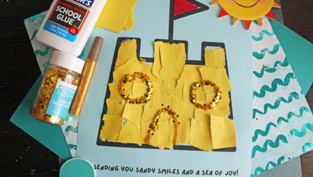 A handmade sandcastle art project made with torn yellow paper and gold glitter on blue paper, with a sun wearing sunglasses in the corner. Elmers glue, glitter, and a craft pen are nearby. Text reads, Sending you sandy smiles and a sea of joy!.
