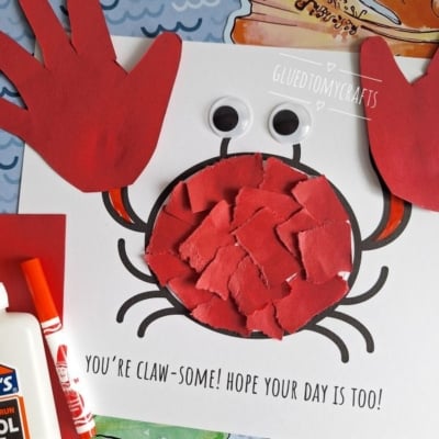 A handmade crab craft made from red paper handprints for claws, a circle filled with red paper pieces for the body, two large googly eyes, and drawn legs on white paper. Text reads You’re claw-some! Hope your day is too!.