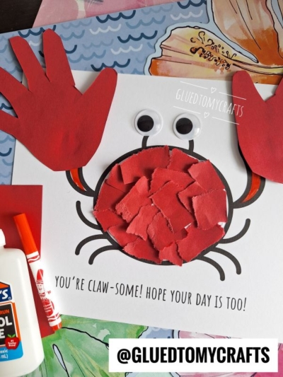 A handmade crab craft made from red paper handprints for claws, a circle filled with red paper pieces for the body, two large googly eyes, and drawn legs on white paper. Text reads You’re claw-some! Hope your day is too!.