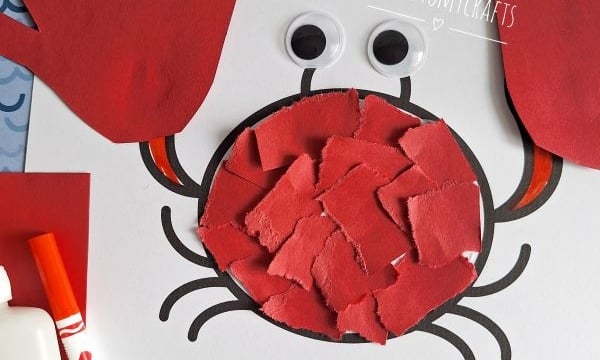 A handmade crab craft made from red paper handprints for claws, a circle filled with red paper pieces for the body, two large googly eyes, and drawn legs on white paper. Text reads You’re claw-some! Hope your day is too!.