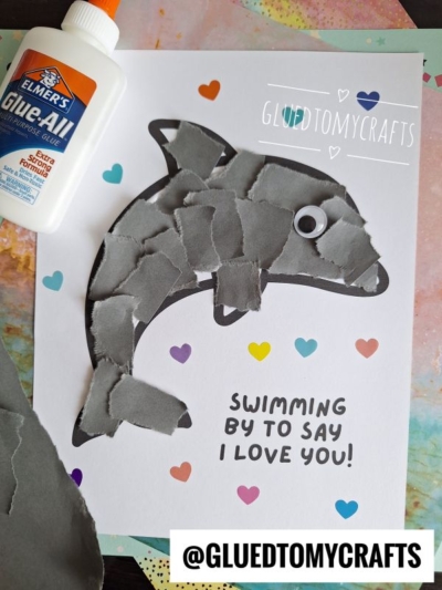 A torn paper dolphin collage with a googly eye is glued onto white paper decorated with colorful hearts and the words Swimming by to say I love you! A bottle of Elmer’s glue and the Instagram handle @GLUEDTOMYCRAFTS are visible.