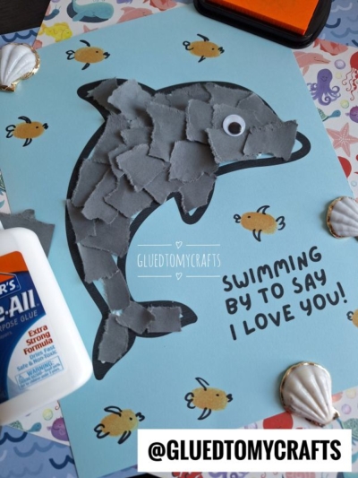 A dolphin made of torn gray paper pieces is glued onto blue paper, surrounded by small fish drawings. Text says “Swimming by to say I love you!” Glue, seashells, and a branded label are also visible.