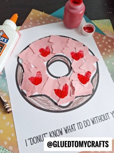 A paper donut craft with pink paper pieces and red painted hearts on a printed donut outline, surrounded by glue, paint, crayon, and colorful paper. Text below reads: I donut know what Id do without you.