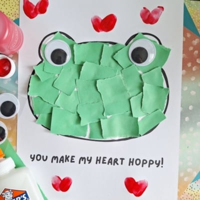 A frog craft with torn green paper pieces pasted inside a frog outline, large googly eyes, red heart-shaped fingerprints at corners, and the text “You make my heart hoppy!” on white paper. Glue and supplies surround it.