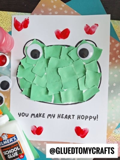 A frog craft with torn green paper pieces pasted inside a frog outline, large googly eyes, red heart-shaped fingerprints at corners, and the text “You make my heart hoppy!” on white paper. Glue and supplies surround it.