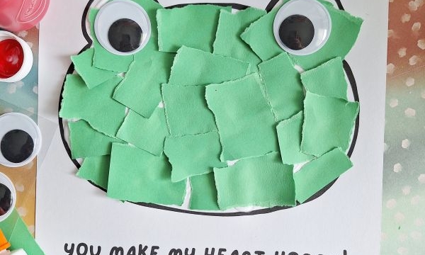 A frog craft with torn green paper pieces pasted inside a frog outline, large googly eyes, red heart-shaped fingerprints at corners, and the text “You make my heart hoppy!” on white paper. Glue and supplies surround it.