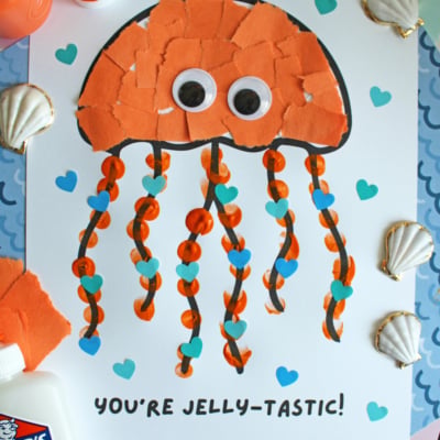 A colorful jellyfish craft made of orange paper pieces, googly eyes, and striped tentacles is on a white sheet with blue hearts. YOURE JELLY-TASTIC! is written below. Seashells, glue, and paint are nearby. “FREE PRINTABLE!!!” is displayed.