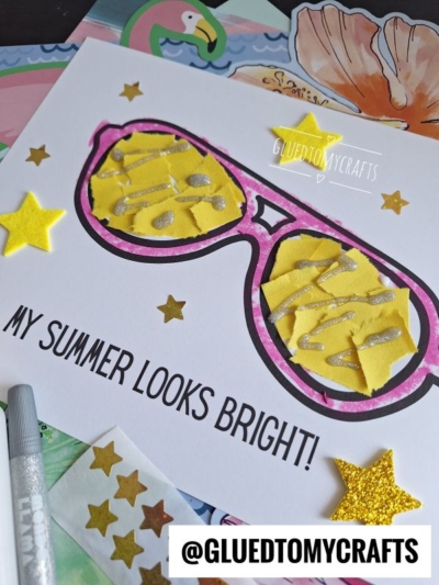 A handmade card features large, colorful sunglasses with yellow lenses decorated with silver glitter. The text reads MY SUMMER LOOKS BRIGHT! Yellow star stickers and craft supplies surround the card.