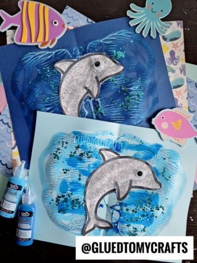 Two dolphin crafts made from paper, glitter, and paint are displayed on colorful patterned backgrounds. Fish and octopus stickers decorate the scene, with @GLUEDTOMYCRAFTS written at the bottom.