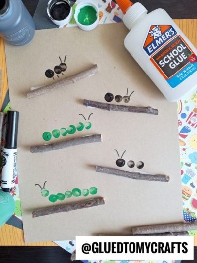 A fun INSECT CRAFT IDEA featuring caterpillars made from green and black paint thumbprints with stick segments, glued to brown paper. Elmer’s glue, paint, and markers are scattered around the playful insect creation.