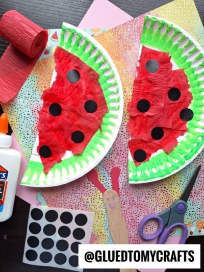 Two paper plates painted and decorated to look like watermelon slices sit on a colorful background. Craft supplies, including glue, scissors, black dot stickers, and red crepe paper, are scattered around.