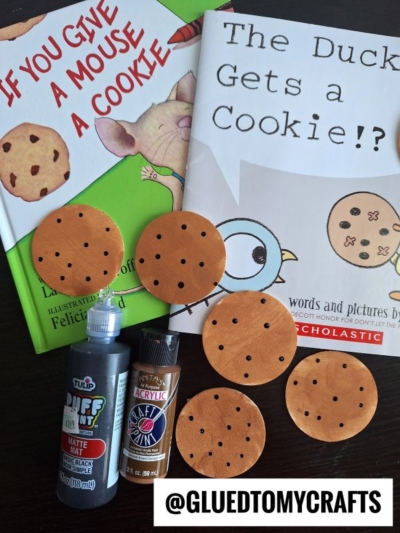 Two children’s books, “If You Give a Mouse a Cookie” and “The Duck Gets a Cookie!?”, are surrounded by craft supplies, paint bottles, and story time sensory bin ideas like fake cookies with chocolate chips on a dark surface. A @GLUEDTOMYCRAFTS label is at the bottom.