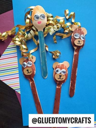 Three handmade puppets made from wooden craft sticks—one with blonde hair and a blue dress, and two brown bear faces with googly eyes—are perfect for summer craft ideas. Decorative paper and gold ribbon add flair. @GLUEDTOMYCRAFTS is at the bottom.