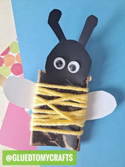 A cute INSECT CRAFT IDEAS project: a bee made from a clothespin wrapped in yellow yarn, with white paper wings, black paper head, googly eyes, and antennae—displayed on blue and multicolored paper. @GLUEDTOMYCRAFTS at the bottom.