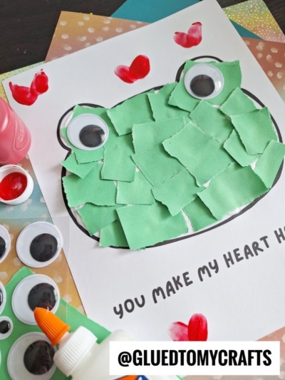 A frog craft made with torn green paper pieces and large googly eyes is glued onto a paper with the words “You make my heart hop.” Craft supplies are scattered around, and the handle @GLUEDTOMYCRAFTS is visible.