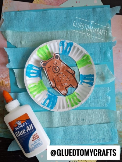 A paper plate with a brown bear drawing is decorated with green and blue paint. Blue crepe paper strips cover the background. A bottle of Elmer’s Glue-All sits on the left. @GLUEDTOMYCRAFTS is written at the bottom.
