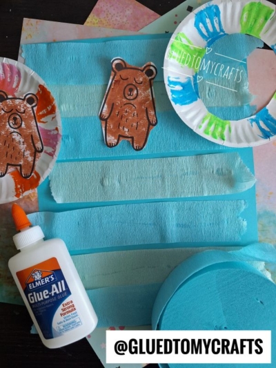 A children’s craft setup with painted paper plates, a bear cutout, blue-striped painted paper, a bottle of Elmer’s Glue-All, and a sponge. The username @GLUEDTOMYCRAFTS is displayed at the bottom.