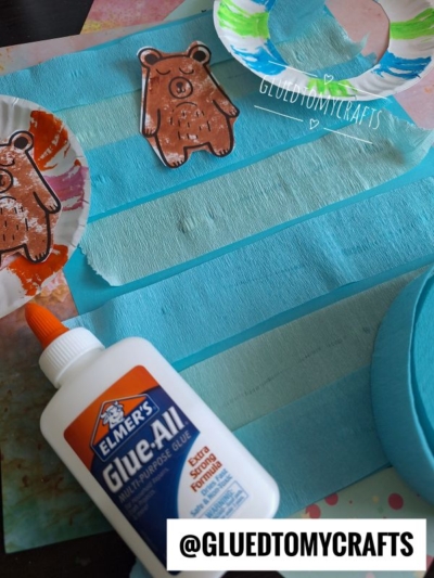 A bottle of Elmer’s Glue-All on a table with blue crepe paper strips, paper bear crafts, and paint. The handle @GLUEDTOMYCRAFTS appears at the bottom of the image.