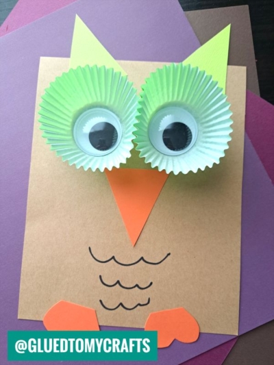 A handmade owl cupcake liner card craft featuring green liners for eyes, large googly eyes, a paper beak, wings, and ears on layered colored paper. The handle @GLUEDTOMYCRAFTS is in the bottom left corner.