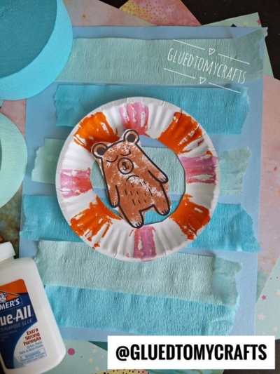 A paper plate painted with orange and pink streaks features a brown bear cutout in the center. It’s placed on a blue textured background with torn paper strips. A bottle of Elmer’s glue and a tag @GLUEDTOMYCRAFTS are visible.
