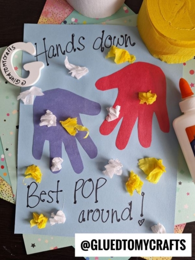 A blue card with red and purple handprints, decorated with crumpled yellow and white tissue paper. Text reads “Hands down, Best POP around!” Glue, craft paper, and tissue are beside the card. @GLUEDTOMYCRAFTS is at the bottom.